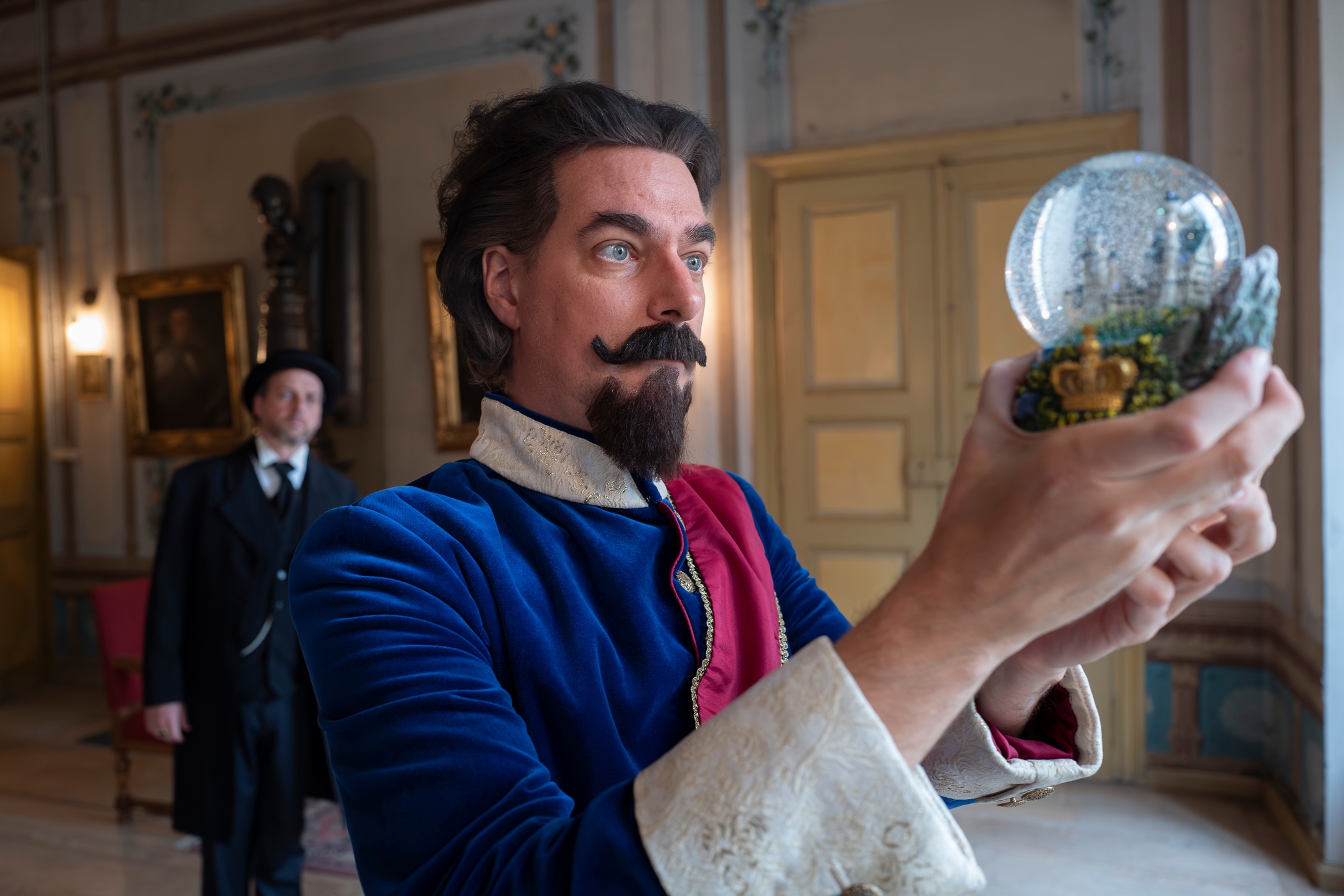Kostümszene: Darsteller als König Ludwig II hält eine Schneekugel, im Hintergrund Dr. Bernhard von Gudden in historischem Interieur