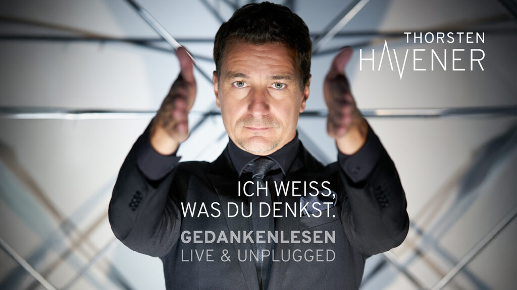 Ein Mann steht frontal zur Kamera und blickt konzentriert nach vorn, die Hände parallel erhoben, als würde er den Betrachter direkt ansprechen. Im Hintergrund geometrische Linien, auf dem Bild der Text „Ich weiß, was du denkst. Gedankenlesen – Live & Unplugged“.