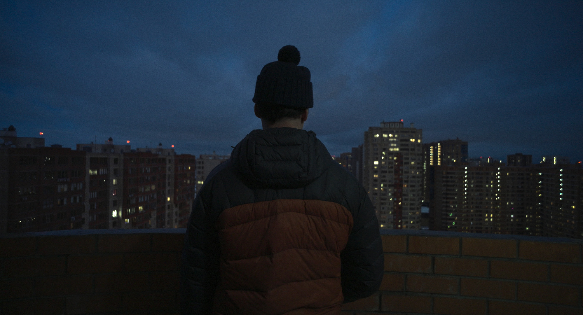 Person mit Winterjacke und Mütze steht auf einem Dach und blickt über eine Stadt mit beleuchteten Hochhäusern in der Abenddämmerung.