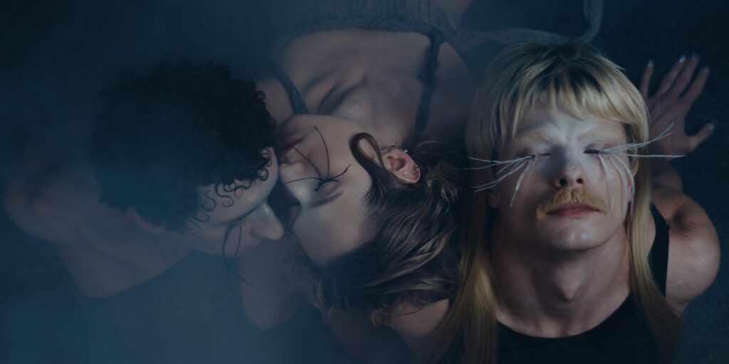 Drei Personen liegen eng beieinander, zwei mit geschlossenen Augen und künstlerischem Make-up, in einer atmosphärisch beleuchteten, inszenierten Szene.
