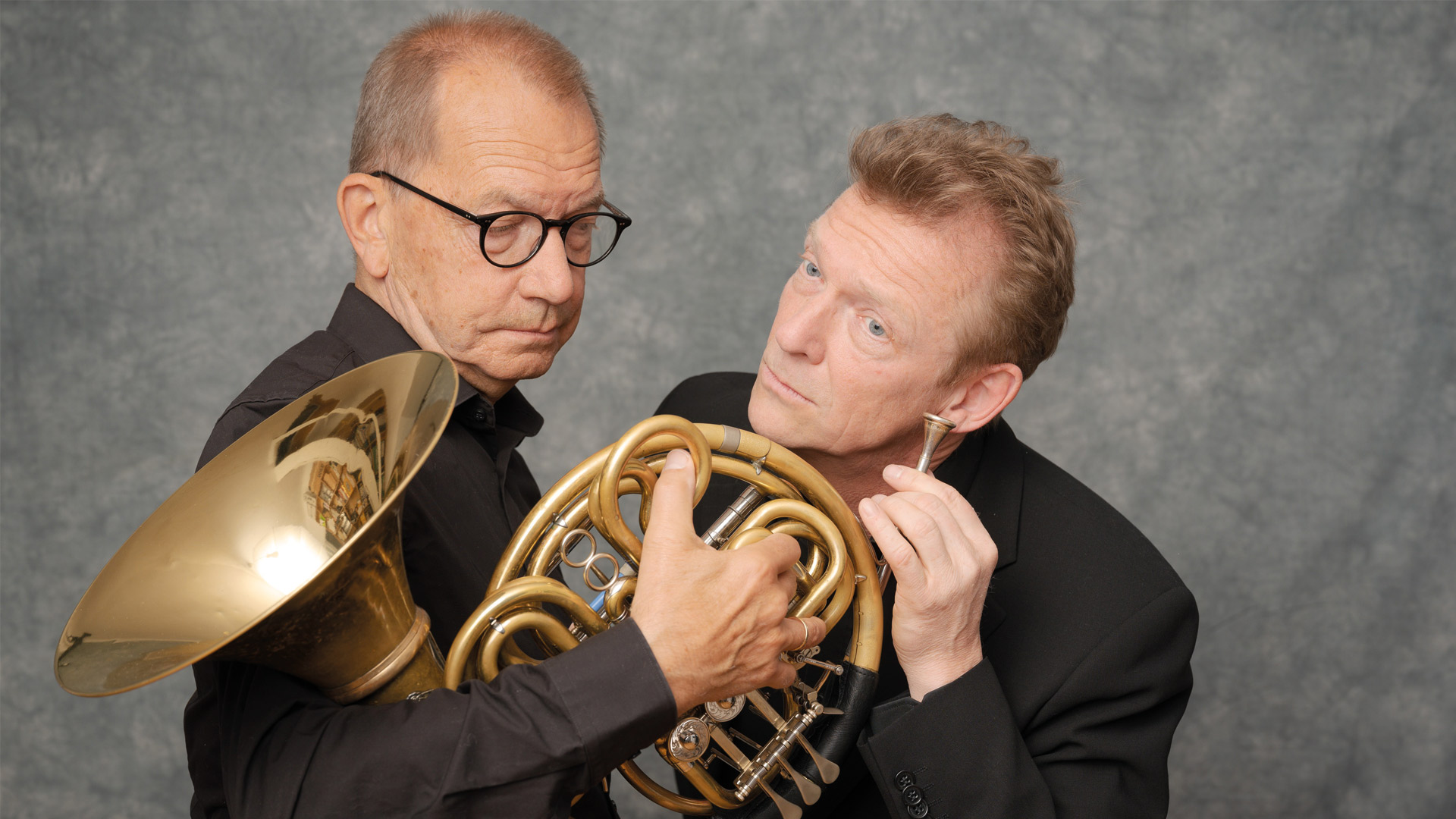 Klaus Wallendorf und Andreas Kowalewitz in schwarzer Kleidung mit einem Waldhorn vor grauem Hintergrund.